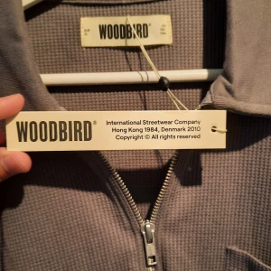 Grå tröja med dragkedja från Woodbird - Säljer en stilren grå tröja från Woodbird. Den har en dragkedja framtill och en praktisk bröstficka. Tröjan är långärmad och har en bekväm passform, perfekt för både vardag och avslappnade tillfällen. Materialet känns mjukt och skönt mot huden.