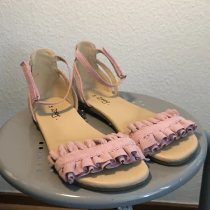 Rosa sandaler med volangdetaljer - Säljer ett par supersöta rosa sandaler från Zoey i storlek 36. De har en fin volangdetalj framtill och justerbara remmar runt ankeln. Perfekta för sommardagar eller en festlig kväll! 🌸