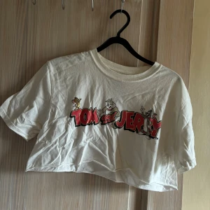 Vit Tom och Jerry t-shirt - Säljer en supercool vit t-shirt med Tom och Jerry-tryck. Den är croppad och har korta ärmar, perfekt för en avslappnad stil. Tillverkad i 100% bomull, vilket gör den både bekväm och lätt att bära. Perfekt för alla som älskar klassiska tecknade serier!