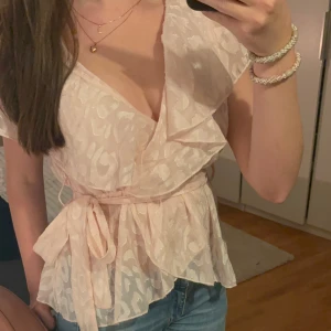 Beige omlottblus från Shein - Säljer en superfin rosa omlottblus från Shein. Den har ett diskret mönster och korta ärmar. Blusen är perfekt för sommaren och har en söt knytning i midjan. Passar både till vardags och fest!