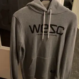 Säljer en snygg och bekväm grå hoodie från WESC. Den har en stor logga på framsidan och en mysig fleece-insida som håller dig varm. Perfekt för både vardag och chill. Den har en klassisk känguruficka och justerbar huva med snören.