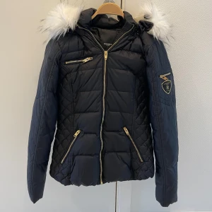 Svart dunjacka med pälskrage - Säljer en snygg svart dunjacka med vit pälskrage. Jackan har en quiltad design och flera dragkedjor i guld. Perfekt för kalla vinterdagar med sin varma och stiliga look. Den har en normal passform och långa ärmar. Märket är R, vilket syns på ärmen.
