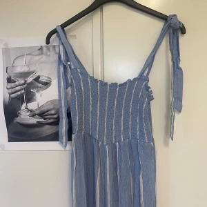 Blå randig jumpsuit från H&M - Säljer en superfin blå jumpsuit från H&M med vita ränder. Klänningen har knytbara axelband och en smockad överdel som ger en bekväm passform. Perfekt för sommardagar eller en avslappnad fest. 🌞