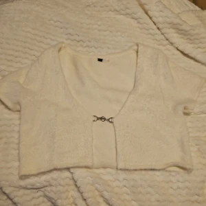 Vit kortärmad bolerotopp - Säljer en superfin vit bolerotopp med korta ärmar. Den är croppad och har en snygg knäppning framtill med en liten metallkedja. Perfekt för att lägga till en extra touch till din outfit. Passar både till vardags och fest! 🕊️