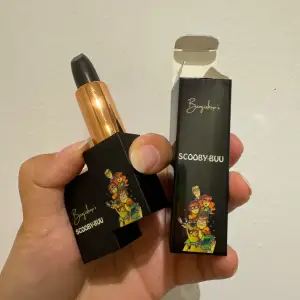 Coolt svart läppstift från Benjiishop med en unik förpackning som har ett Scooby-Doo-tema. Läppstiftet har en elegant guldig hylsa och kommer i en snygg svart kartong med färgglada illustrationer. Perfekt för en djärv look!