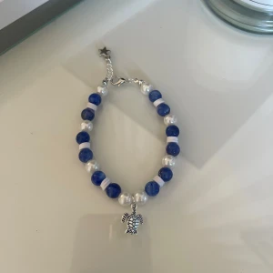 Blått och vitt pärlarmband med sköldpaddshänge - Säljer ett supergulligt armband med blå och vita pärlor. Armbandet har ett sött sköldpaddshänge och en stjärnformad detalj vid låset. Perfekt för att ge en färgklick till din outfit!