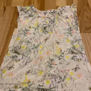 Säljer en söt blommig topp från H&M i storlek 122. Toppen är vit med ett färgglatt blommönster i rosa, gult och grönt, samt små bin och fjärilar. Den har en lös passform och är ärmlös, perfekt för varma sommardagar. Bra skick och redo för ny ägare!