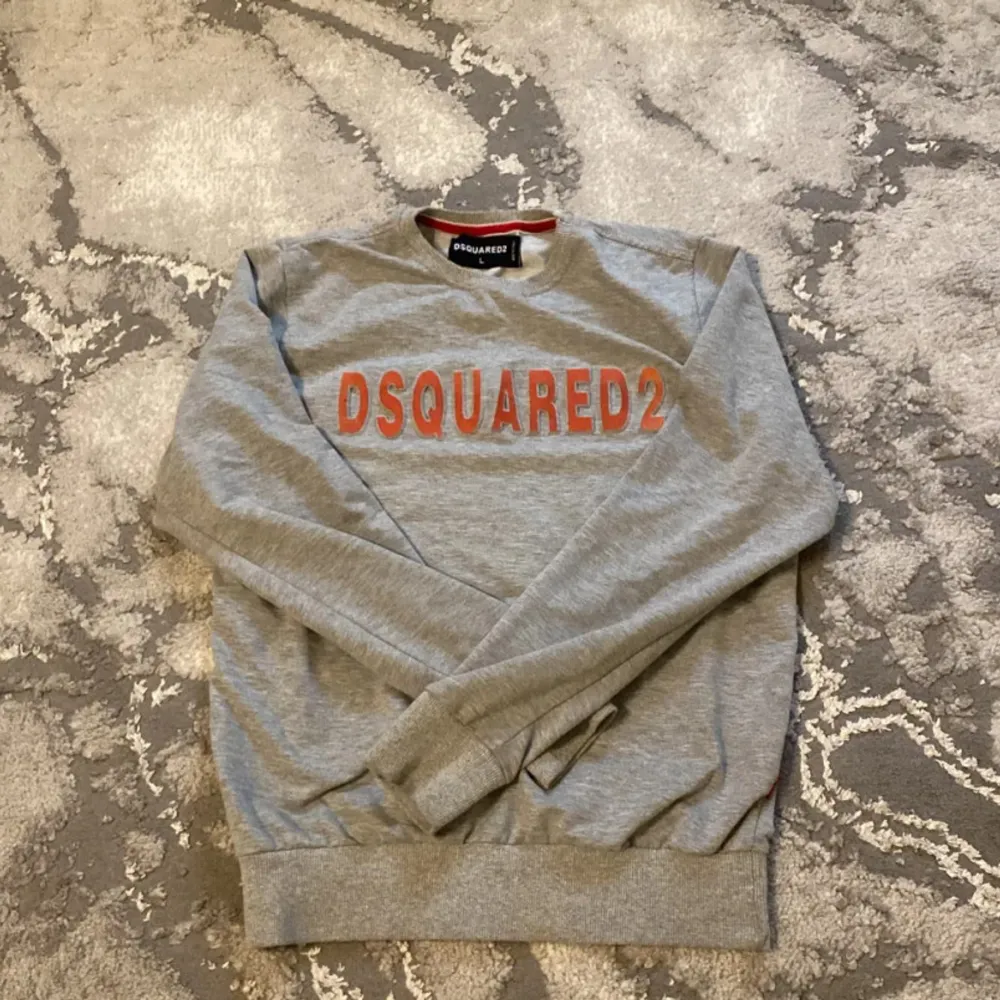 Säljer en snygg grå sweatshirt från DSQUARED2 i storlek L. Tröjan har ett coolt rött tryck på framsidan med märkesnamnet. Den är perfekt för en avslappnad stil och passar bra till både jeans och joggers. Materialet känns mjukt och skönt mot huden. Perfekt för höst och vår!. Tröjor & Koftor.