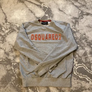 Grå sweatshirt från DSQUARED2 - Säljer en snygg grå sweatshirt från DSQUARED2 i storlek L. Tröjan har ett coolt rött tryck på framsidan med märkesnamnet. Den är perfekt för en avslappnad stil och passar bra till både jeans och joggers. Materialet känns mjukt och skönt mot huden. Perfekt för höst och vår!