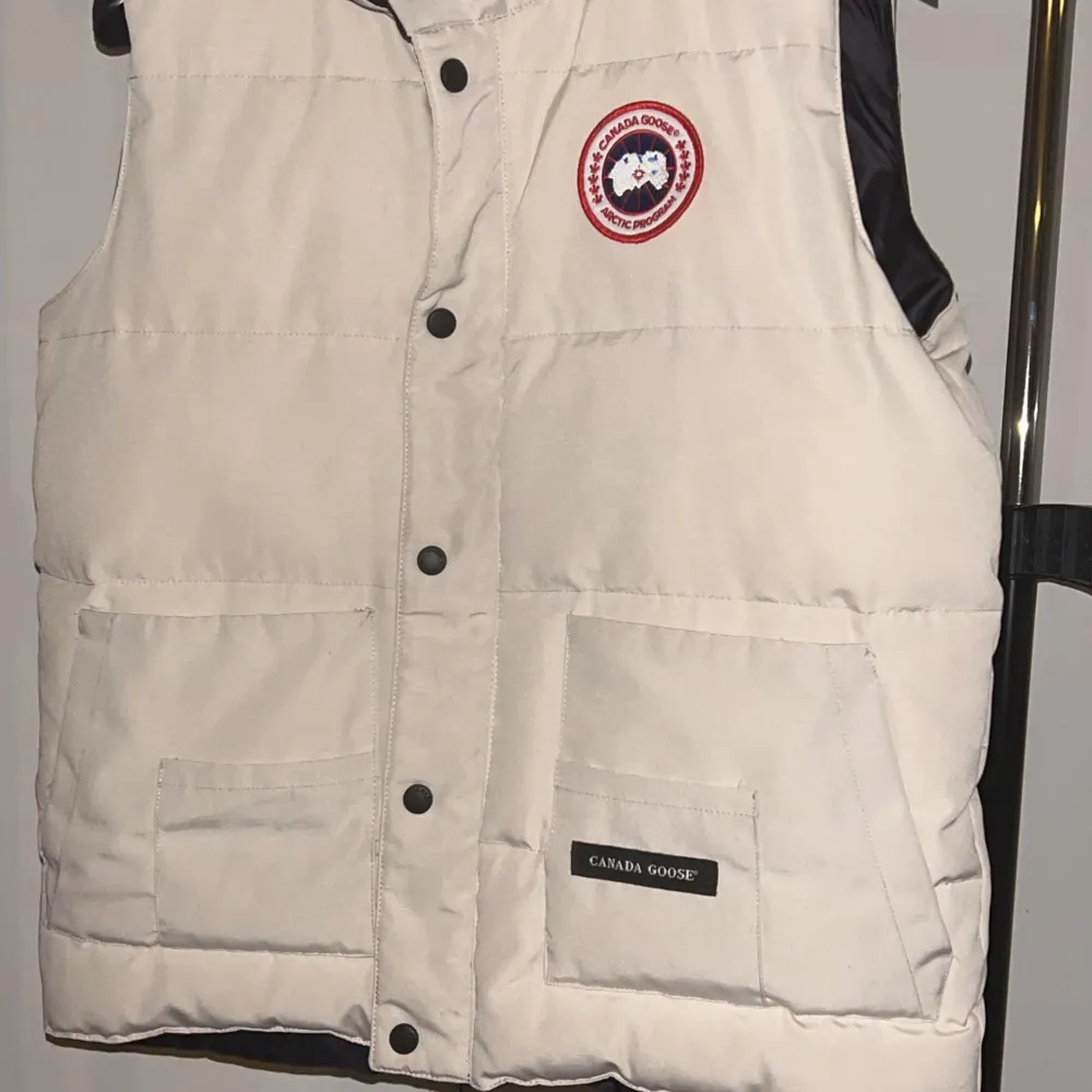 Säljer en snygg och varm beige dunväst från Canada Goose. Västen har en klassisk design med knappar framtill och två stora fickor. Perfekt för kyliga dagar och passar bra till både vardag och utomhusaktiviteter. Märket är välkänt för sin kvalitet och hållbarhet. Passa på att köpa denna stilrena väst!. Jackor.