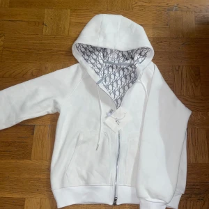 Vit hoodie med mönstrad huva - Säljer en stilren vit hoodie med dragkedja och en mönstrad insida i huvan. Perfekt för en avslappnad look. Huvan har ett snyggt grått mönster som ger en extra touch. Pris kan diskuteras vid snabb affär!!!