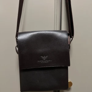 Brun axelväska från Giorgio Armani - Snygg brun axelväska från Giorgio Armani i skinn. Väskan har en justerbar axelrem och en praktisk dragkedja på baksidan. Perfekt för att bära dina nödvändigheter med stil.