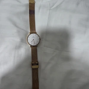 Guldfärgad klocka från Daniel Wellington - Elegant guldfärgad klocka från Daniel Wellington med vit urtavla och minimalistisk design. Klockan har ett mesh-armband i guld och en stilren look som passar perfekt för alla tillfällen.