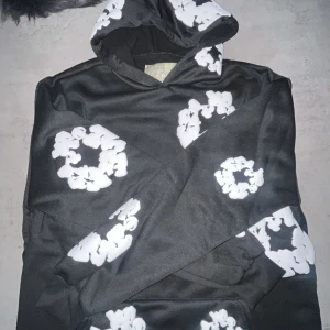 Svart hoodie med blommönster från Denim Tears - Säljer en svart hoodie från Denim Tears med ett unikt vitt blommönster. Hoodien har en klassisk design med huva och en stor ficka framtill. Perfekt för en avslappnad stil.