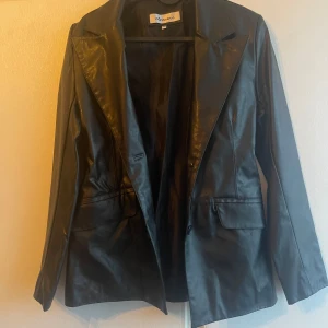 Svart skinnjacka från Dreams - Snygg svart skinnjacka från Dreams med klassisk design. Jackan har långa ärmar och en stilren look, perfekt för en cool och elegant stil. Tillverkad i 80% PVC och 20% polyester med foder i 100% polyester.