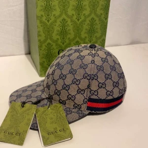 Beige keps från Gucci med GG-mönster - Snygg beige keps från Gucci med det ikoniska GG-mönstret. Kepsen har en röd och blå randdetalj på sidan och kommer i originalförpackning. Perfekt för att ge din outfit en lyxig touch.