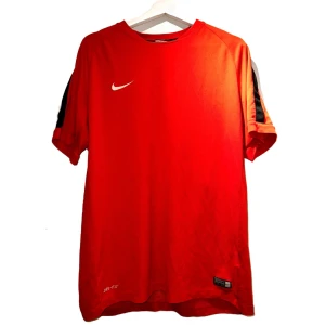 Röd Dri-FIT t-shirt från Nike - Snygg röd Dri-FIT t-shirt från Nike med korta ärmar och svart detalj på axlarna. Perfekt för träning eller en aktiv dag. Den har en klassisk Nike-logga på bröstet.