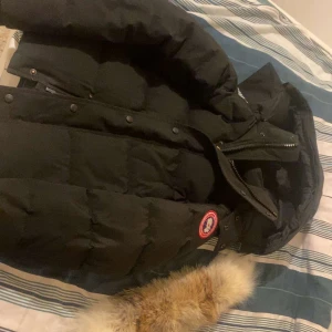 Svart dunjacka med pälsdetalj - Säljer en svart dunjacka från Canada Goose med avtagbar pälsdetalj på huvan. Jackan har dragkedja och knappar framtill samt en broderad logotyp på ärmen. Perfekt för kalla vinterdagar.