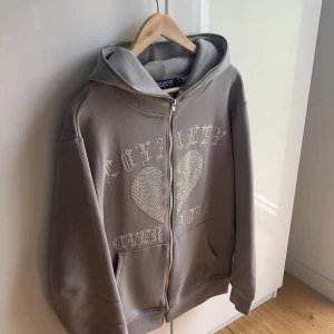 Rhinestone zip Hoodie  - Säljer en beige hoodie från Trotherz med dragkedja och ett glittrigt hjärtmönster på framsidan. Använd ett fåtal par gånger, aldrig tvättad alla nitar sitter som om de vore helt nya. Finns bevis på att den direkt är köp från hemsidan.🤩Kvitto finns! / bevis medföljs.  Mvh ItsForFits 🌟