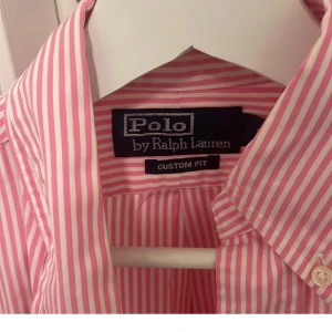 Rosa och vit randig skjorta från Polo Ralph Lauren - Snygg rosa och vit randig skjorta från Polo Ralph Lauren med broderad logga i blått på bröstet. Skjortan har långa ärmar och knappar framtill. Perfekt för en stilren look.