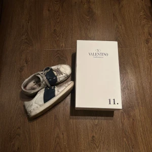Valentino Garavani sneakers - Säljer ett par Valentino Garavani sneakers i vitt med blå detaljer. Skorna är i använt skick som ni kan se på bilderna därav priset. Allt OG finns kvar. Storlek 43 passar 44 också. Riktigt snygga skor och passar till vilken outfit som helst. Bara kosmetiskt slitna men inga hål etc 