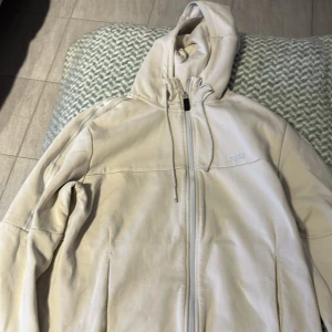 Beige hoodie från Boss - Säljer en stilren beige jacka från Boss med huva och dragkedja. Perfekt för en avslappnad look. Jackan har en enkel design med diskret logga på bröstet.