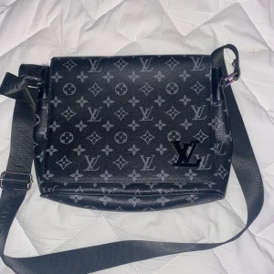 Svart axelväska  - Snygg svart axelväska med justerbar rem och monogrammönster. Väskan har en praktisk design med lock och är perfekt för att bära dina dagliga nödvändigheter. Den har en stilren look som passar till många olika outfits.