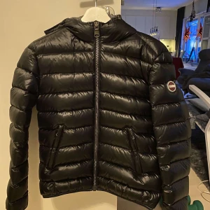 Svart pufferjacka från Colmar - Snygg svart pufferjacka från Colmar med huva och dragkedja framtill. Jackan har en quiltad design och är perfekt för kyligare väder. Den har en normal passform och långa ärmar. Storlek 8 USA vilket motsvarar ungefär 38.