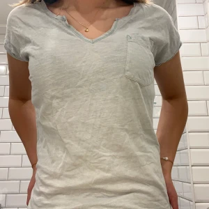 Ljusblå t-shirt med bröstficka - Säljer en ljusgrå t-shirt med v-ringning och en praktisk bröstficka. T-shirten har korta ärmar och är Perfekt för en avslappnad stil. Lite urtvättad men svagt blå ändå. Skriv privat för fler bilder 🩷