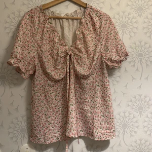 Blommig blus med knytning - Charmig blommig blus i rosa och vit med korta ärmar och knytning framtill. Perfekt för en somrig look med volangdetaljer vid ärmslut och halsringning.