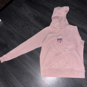 Rosa hoodie från Gant - Säljer en snygg rosa hoodie från Gant med ett broderat emblem på bröstet. Tröjan har en klassisk design med långa ärmar och en praktisk ficka framtill. Perfekt för en avslappnad stil. Har även 2 små blå prickar på ärmen 