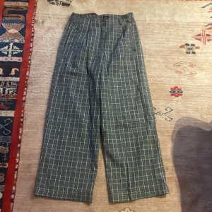 Gröna rutiga byxor - Snygga gul gråa byxor med vitt rutmönster. De har en vid passform och är perfekta för en stilren look. Byxorna har en klassisk design med knapp och dragkedja framtill.