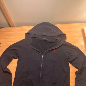 Mörkblå hoodie från Polo Ralph Lauren - Säljer en snygg mörkblå hoodie från Polo Ralph Lauren med dragkedja och den klassiska röda loggan broderad på bröstet. Perfekt för en avslappnad stil med en skön huva och långa ärmar. Passar bra till både vardag och helger det är storlek 165
