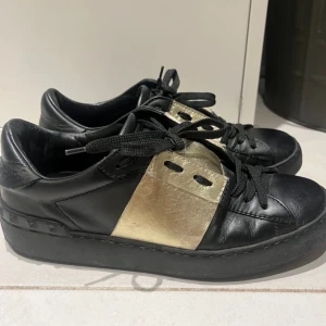 Svarta sneakers med gulddetaljer - Snygga svarta sneakers med gulddetaljer och snörning. Skorna har en modern design med en bred guldfärgad rem över ovansidan. Perfekta för att ge en stilren touch till din outfit.