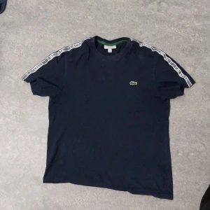 Mörkblå t-shirt från Lacoste - Säljer en mörkblå t-shirt från Lacoste med korta ärmar och vita detaljer längs axlarna. Den har den klassiska krokodilloggan broderad på bröstet. Perfekt för en avslappnad stil.