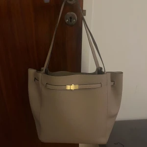 Beige axelväska med gulddetalj - Snygg beige axelväska i syntetmaterial med en elegant gulddetalj framtill. Väskan har en rymlig insida med en praktisk innerficka med dragkedja för extra förvaring. Perfekt för att bära med sig alla nödvändigheter med stil.