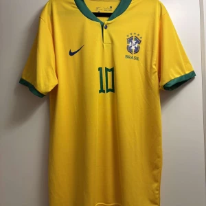Gul Brasilien fotbollströja från Nike - Säljer en gul Brasilien fotbollströja från Nike med gröna detaljer. Tröjan har korta ärmar och nummer 10 på framsidan samt Neymar Jr. på baksidan. Perfekt för fotbollsfans!