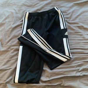 Svarta träningsbyxor från Adidas - Snygga svarta träningsbyxor från Adidas med klassiska vita ränder längs sidorna. Byxorna har elastisk midja och dragkedja vid bensluten för extra komfort. Perfekta för träning eller en avslappnad stil.