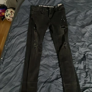 Svarta jeans från Black Squad Projects - Snygga svarta jeans med slitna detaljer och färgstänk från Black Squad Projects. De har en tight passform och är perfekta för en edgy stil. Midjan är låg och byxorna har en klassisk femficksdesign.