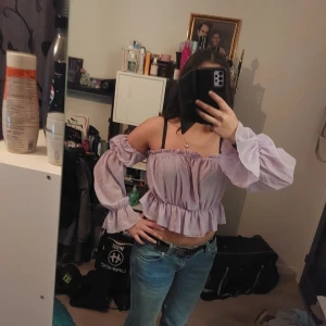 Ljuslila offshoulder blus - Säljer en söt ljuslila offshoulder blus med volangdetaljer. Blusen har långa ärmar och en croppad passform, perfekt för en somrig look. Den är tillverkad i ett lätt och luftigt material.