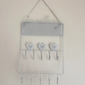 Vit smyckeshängare med hjärtan - Charmig vit smyckeshängare med dekorativa hjärtan och flera krokar för att organisera dina smycken. Perfekt för att hänga upp halsband och armband. Hängaren har en fin, perforerad design upptill och ordet 'jewellery' i metalltråd.