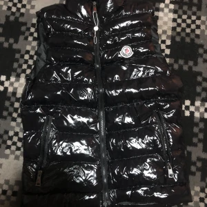 Svart glansig dunväst från moncler  - Snygg svart glansig dunväst från Moncler med dragkedja och logotyp på bröstet. Perfekt för kyliga dagar när du vill hålla stilen. Västen har praktiska fickor med dragkedjor.