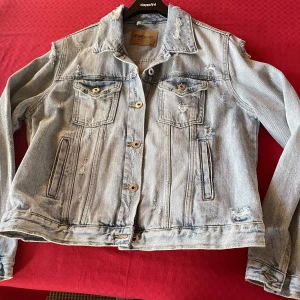 Jeansjacka med hål här och där  - Jeansjacka, ”trasig” både här och där  Materialet är 100% bomull Använd ett fåtal gånger 60 cm lång 52 cm tvärs över byst , mätt från ärm till ärm Storleksmärkt XL 2 bröstfickor med ficklock  2sidofickor 