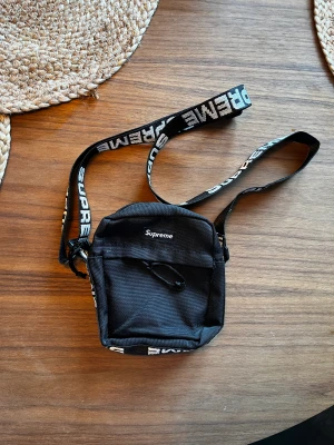 Supreme SS18 Axelväska - Supreme axelväska i väldigt bra skick förrutom 1 flag ett litet jack ovanför dragkedjan men inget man ser. (Kan skicka bild på det)  Materialet är reflex vid justerbara bandet och den har 2 fack, ett stort och ett litet båda med dragkedja.