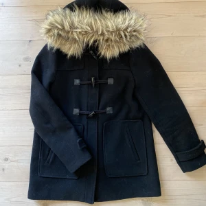 Kappa med ullblandninh och fejkpäls - Kappa i duffel modell i ylleblandning. Luva med avtagbar fejkpäls. Dragkedja och duffelknappar. Inköpt för ca 5-6 år sedan, men sparsamt använd. 