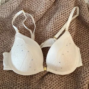 Bh från Marie Meili - Superfin vit bh med spetsdetaljer och små stenar. Man knäpper den fram med den guldiga delen. Storlek 80A med lite vaddering i kupan. Det följer med en del man in fästa på banden där bak. Helt oanvänd. 