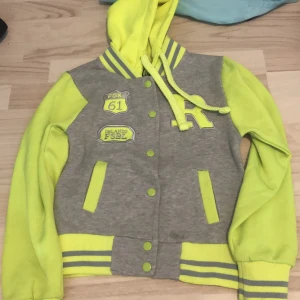 Huvtröja - Grå huvtröja med neon gula ärmar,huva och detaljer  Mitten knappen samt den näst längst ner funkar inte, annars fint skick 