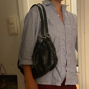 Trendig skitsnygg svart axelväska  - Trendig axelväska med rött (!!) innertyg och två innerfickor. Båda dragkjedjorna är i gott skick. Ej äkta läder och lite slitage har uppkommit när jag haft för tung last, men det syns knappt och går nog att sy igen med några stygn för hand! Går att använda till allt från skola till gym osv!