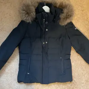 Woolrich jacka i kort modell, mörkblå, avtagbar luva och pälskrage. Matt utsida. Varm! Sparsamt använd, dock har en söm i fodret gått upp (se bild), påverkar ej funktionen och som nog är enkelt att fixa.  Bra skick! Säljes pga för liten.  Tts 