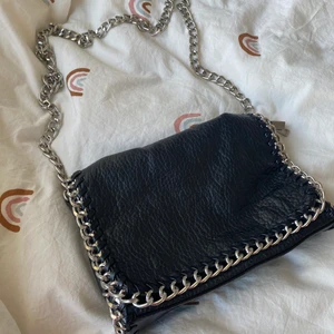 Tiamo väska, svart - Trendig svart axelremsväska i skinnimitation med kantad sileverfärgad länk. Ett fack med dragkedja på baksidan och ett fack med dragkedja på insidan. Axellänk 105 cm. Längd 28cm och  höjd 18 cm. Har använt fåtal gånger. 